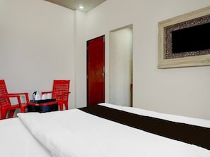 A hotel room with a bed, two red chairs, a small table with a kettle and cups, and a mirror on the wall.