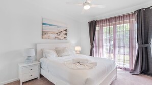 Un dormitorio con cama, mesitas de noche, ventilador de techo, ventana con cortinas y un cuadro en la pared.