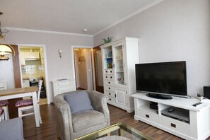 Ein Wohnzimmer mit Fernseher, einer Couch, einem Esstisch und einer Küche im Hintergrund.
