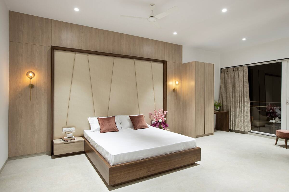 A modern bedroom with a large bed, wooden headboard, bedside lamps, and a sliding glass door to the balcony.
