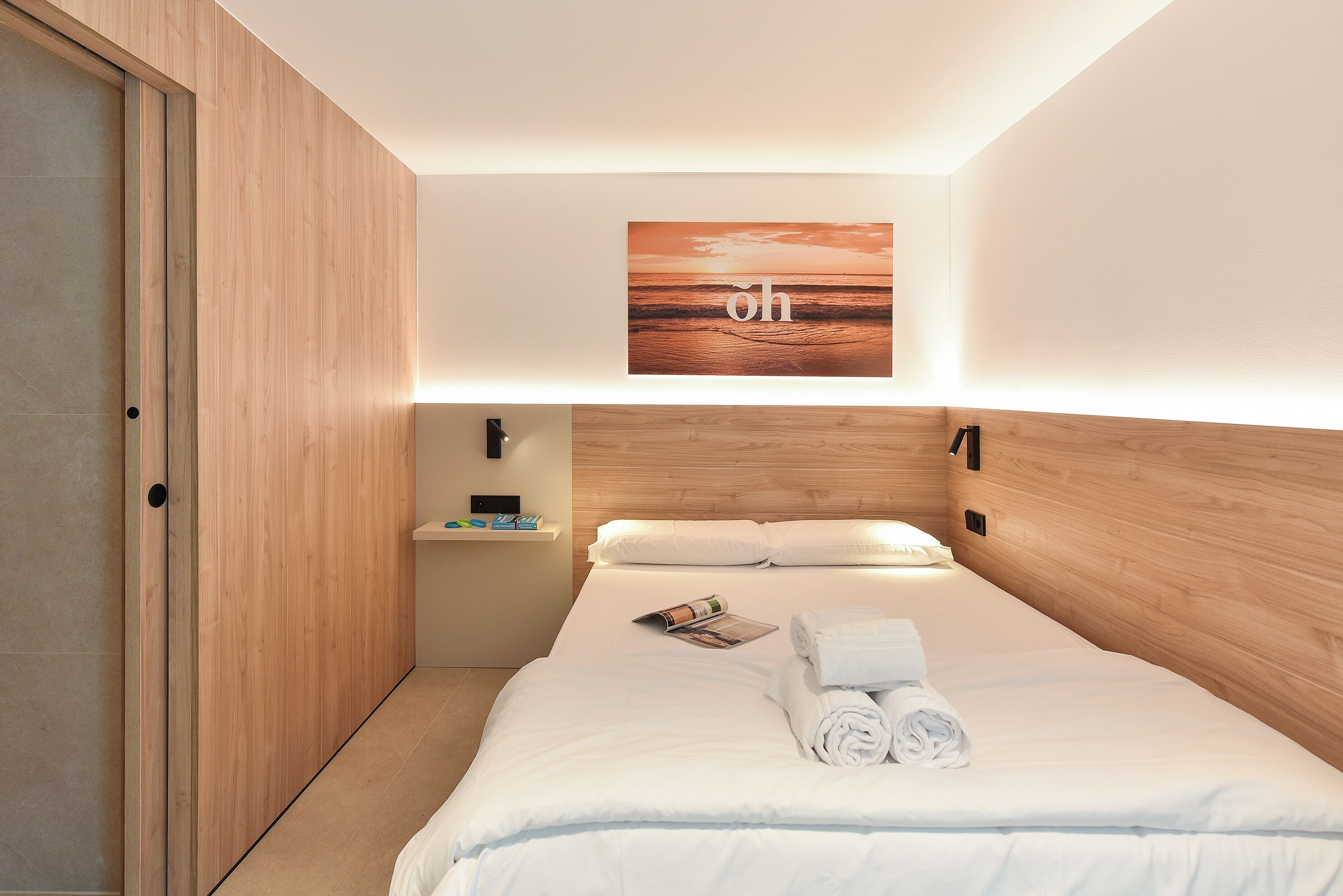 Habitación de hotel con cabecera de madera, cama bien tendida con ropa blanca, una obra de arte colgada en la pared y un estante pequeño con artículos de aseo.