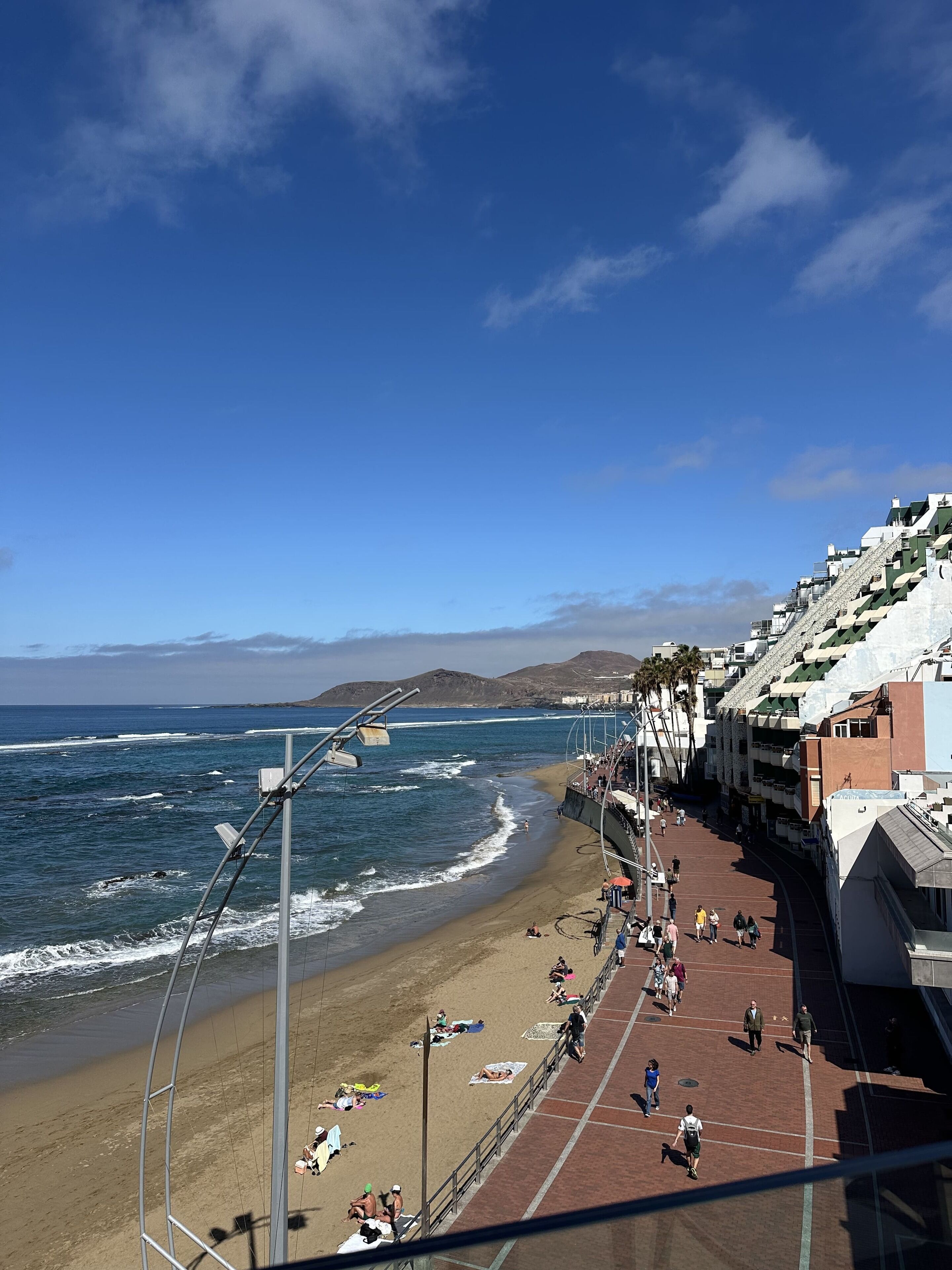 Une promenade en bord de mer, avec une plage, l’océan et une colline parsemée de bâtiments.