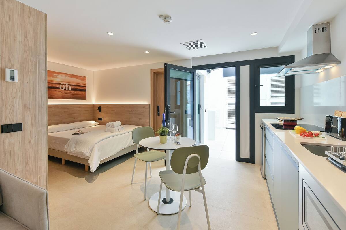 A modern hotel room with a bed, a dining table with chairs, a kitchen area, and a large window.