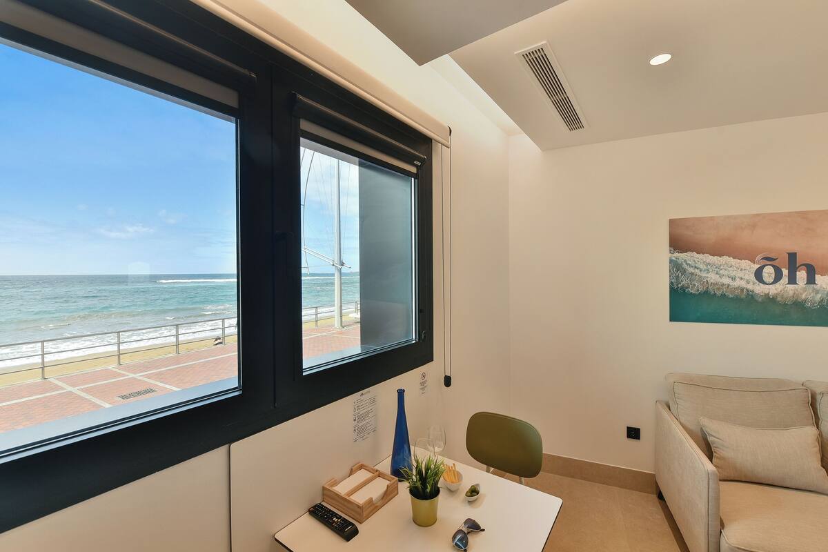 A room with a large window overlooking a beach, a sofa, a small table with a remote control, a vase, and a painting on the wall.