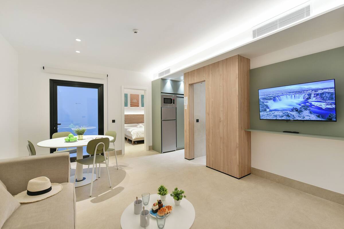 A modern hotel room with a dining area, kitchenette, and a large TV screen displaying a waterfall.