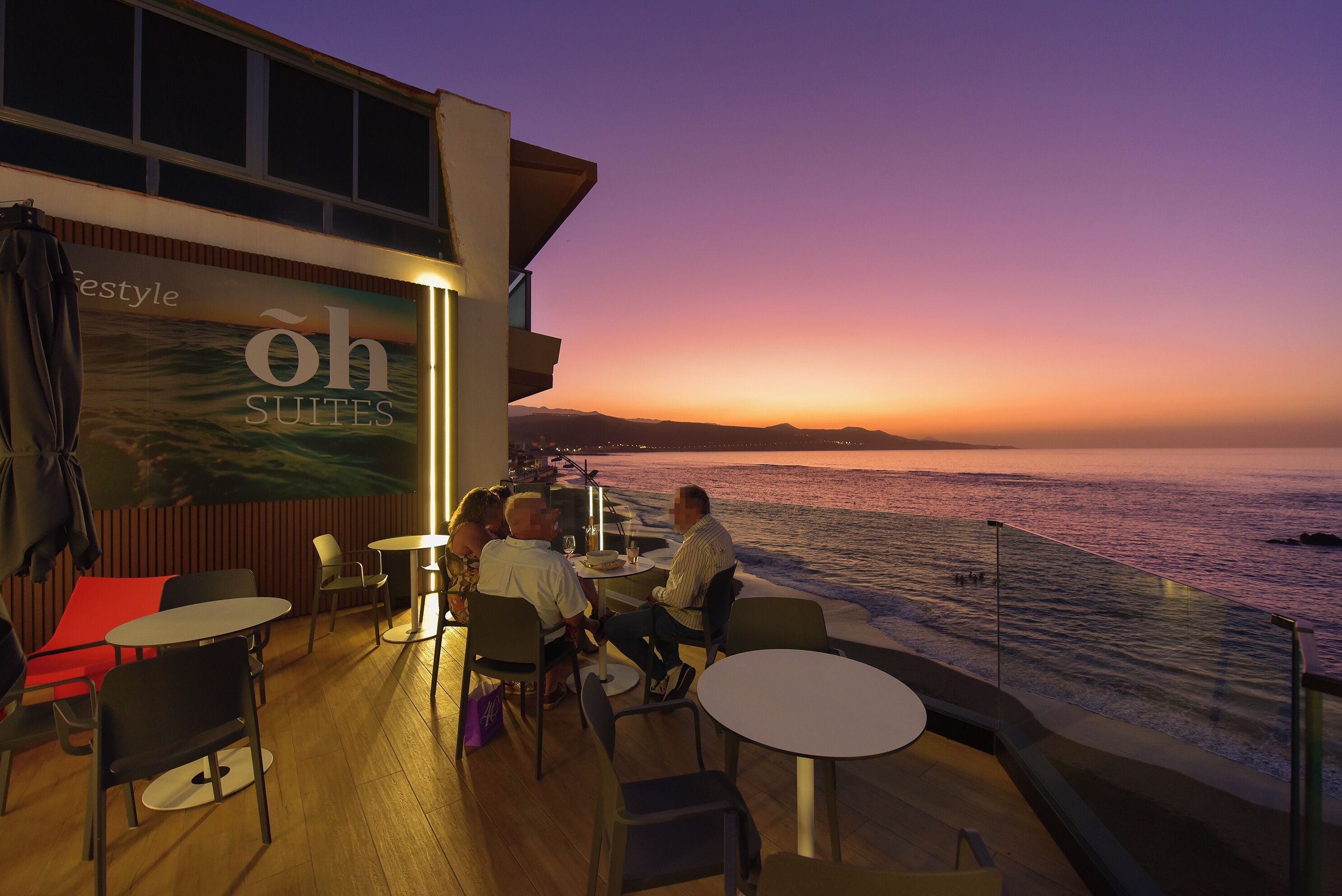 A dining area with tables and chairs, a view of the ocean, and a sign for 'oh SUITES'.