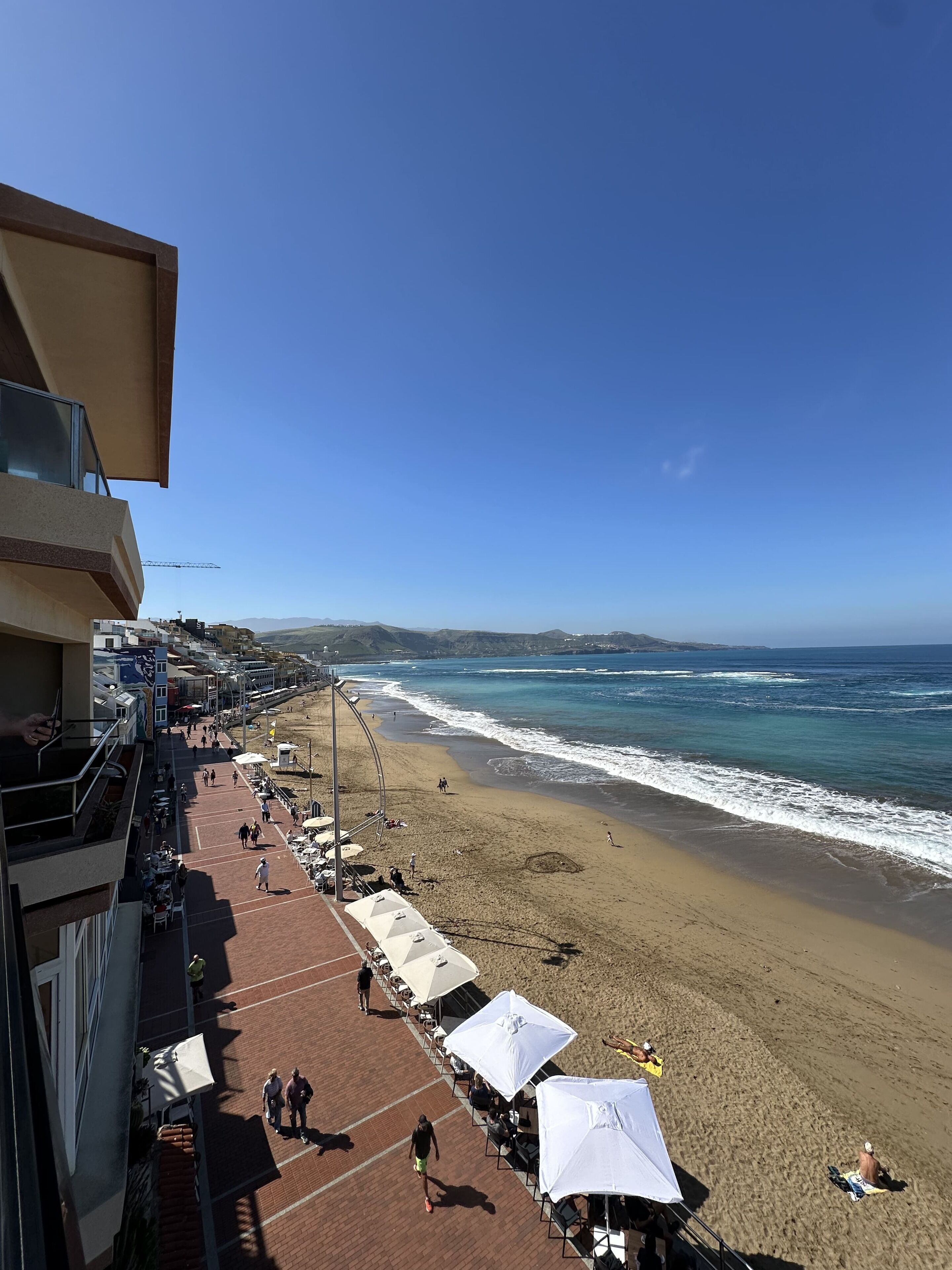 A beachside promenade with beach umbrellas, a clear view of the ocean, and a distant mountain range.