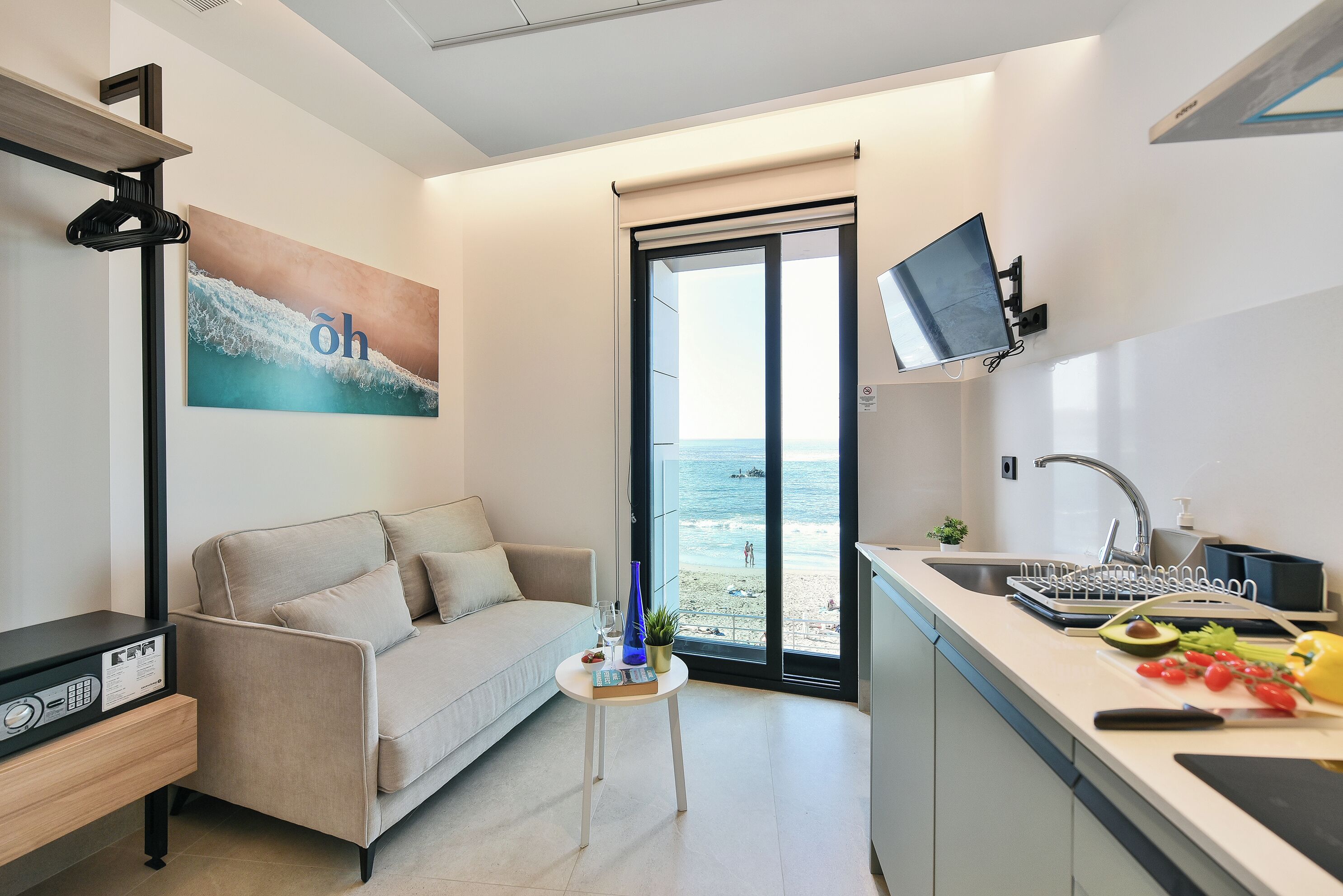 Et moderne værelse med sofa, et lille bord med et planter, et tv ophængt på væggen og udsigt til stranden gennem en skydedør.
