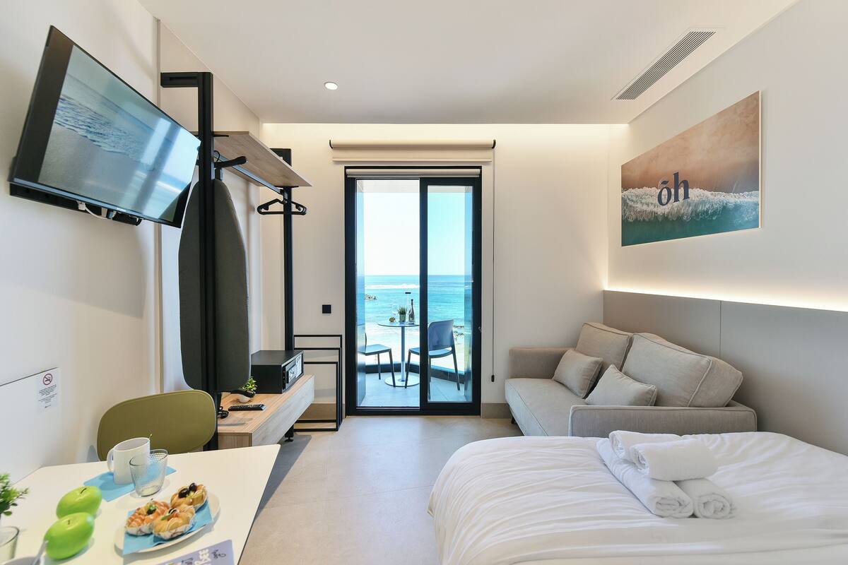 A hotel room with a bed, a sofa, a desk with a computer, a TV, and a view of the sea through a sliding door.