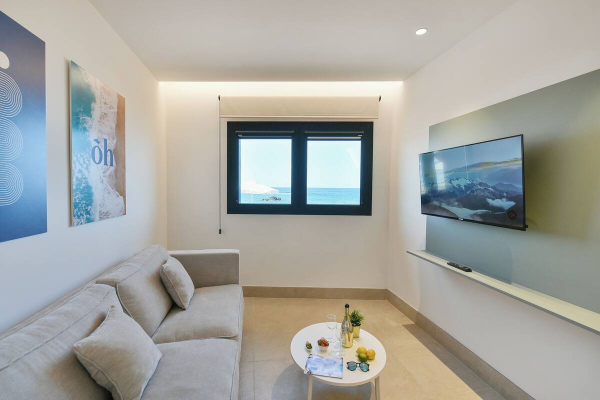 A modern living room with a beige sofa, a round coffee table with drinks and a remote control, a TV on the wall, and a window with a view of the sea.