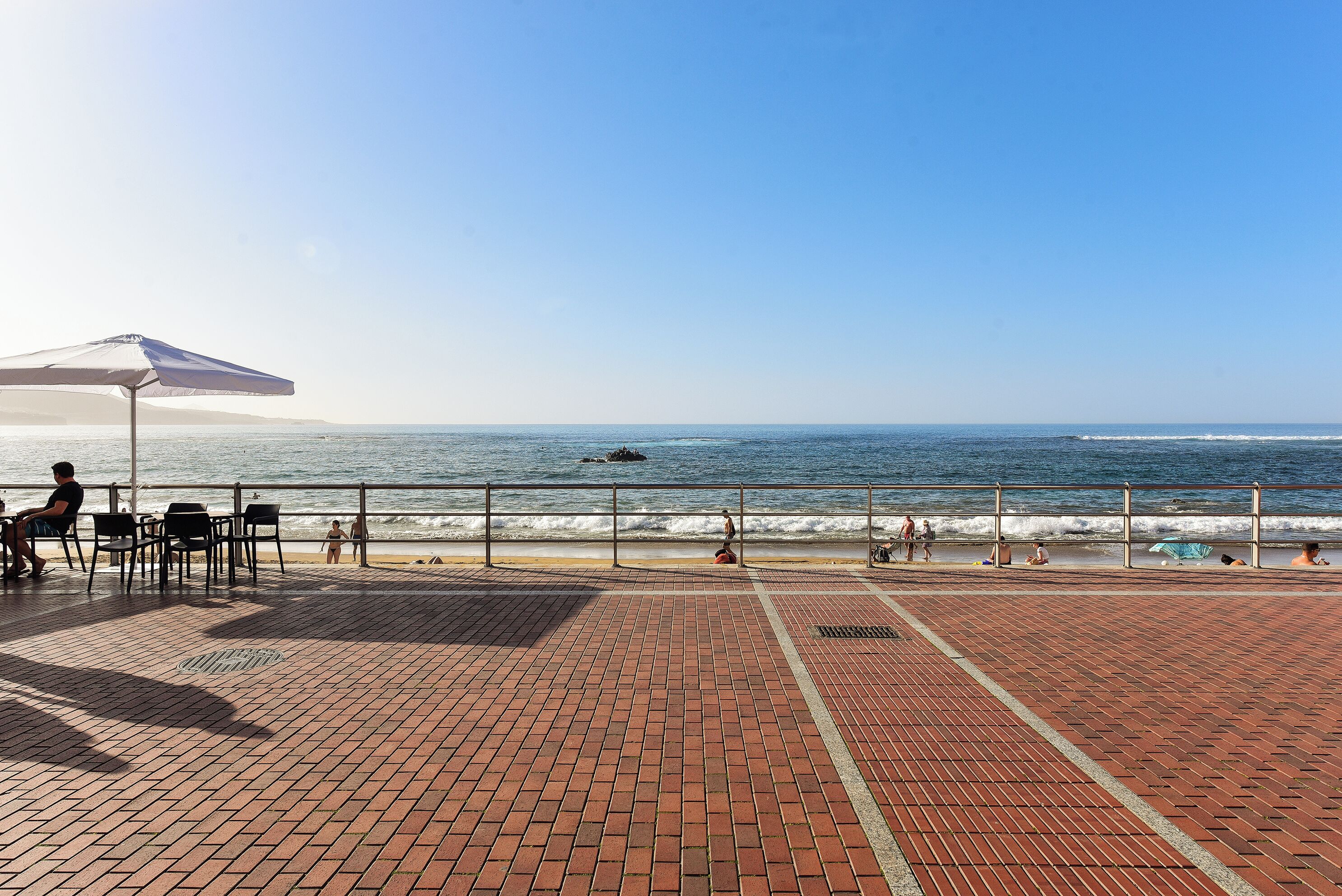 A beachside patio with brick flooring, a white umbrella, and a railing overlooking the ocean.