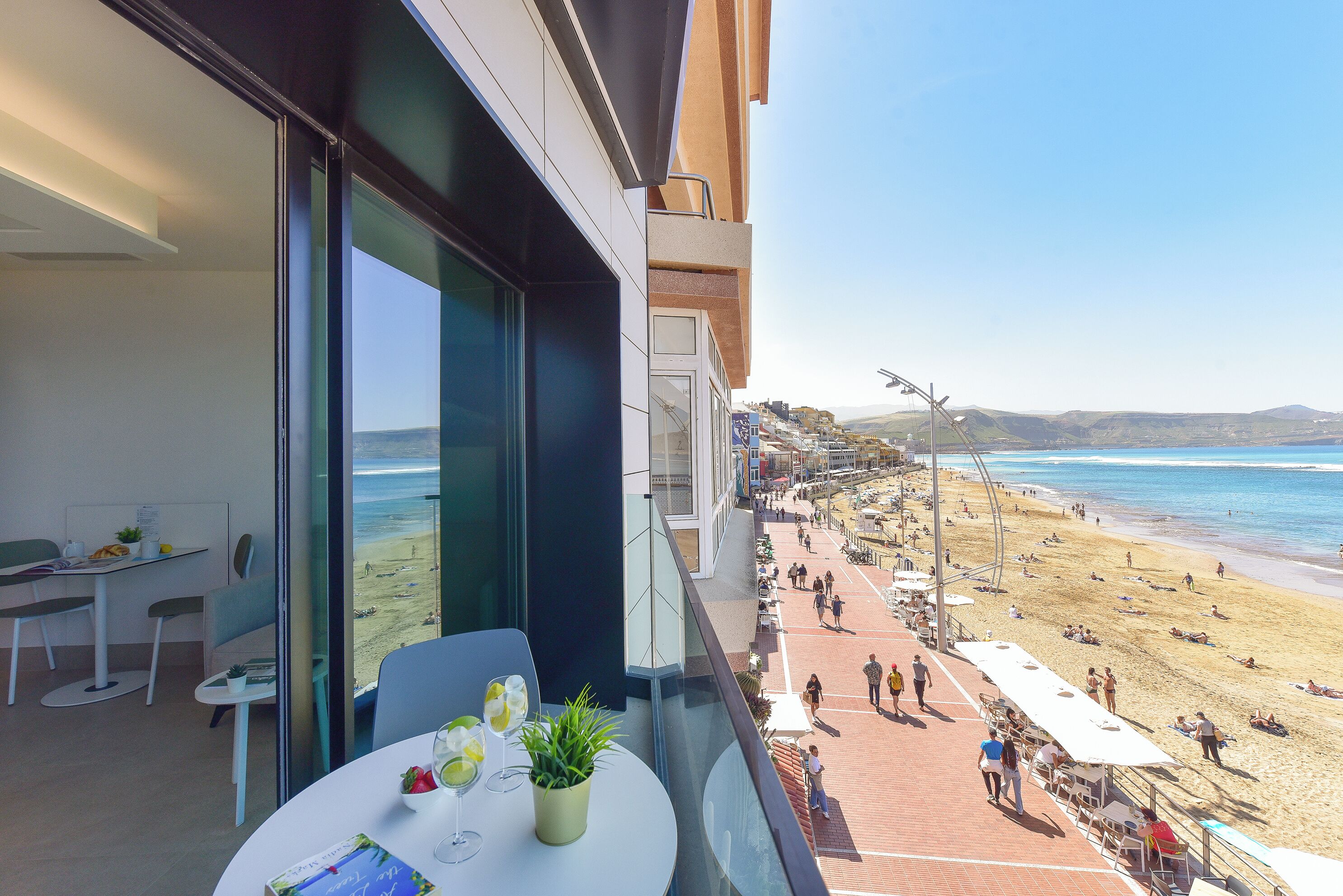 Un balcón con una mesa lista para dos personas, con vista a una playa con gente y sombrillas.