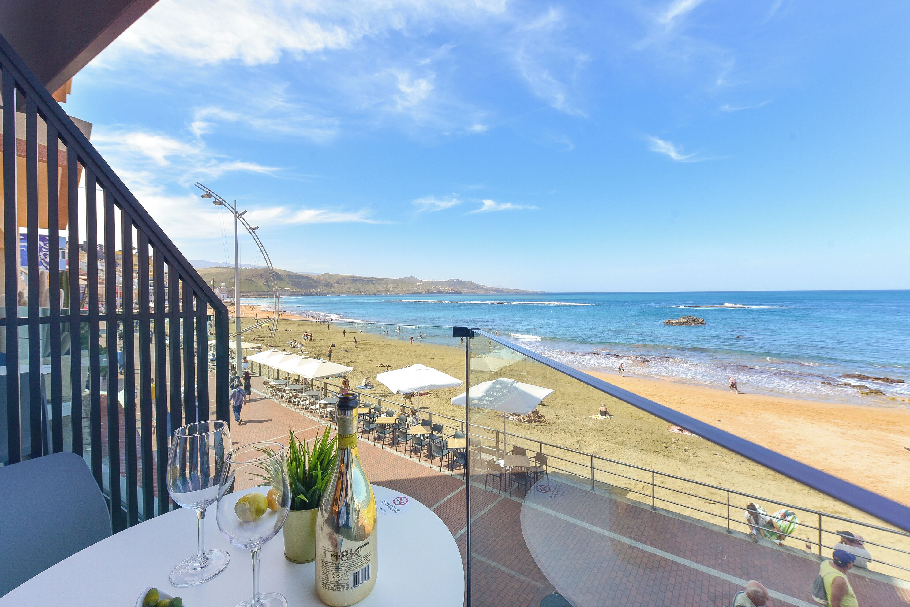 A balcony with a table set for two, overlooking a beach with umbrellas and people.