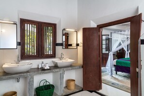 Ein Badezimmer mit zwei Waschbecken, einem großen Spiegel und einer Holztür, die in ein Schlafzimmer mit Bett und Baldachin führt.