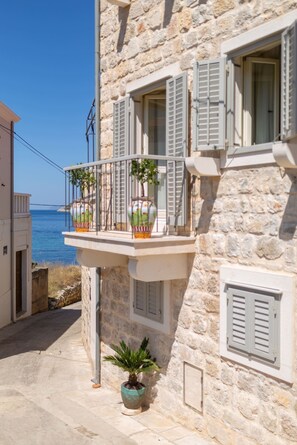 Un edificio de piedra con balcón que da al mar, con plantas en macetas y contraventanas.