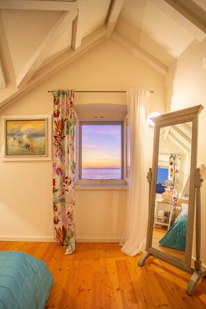 A bedroom with a wooden floor, a window with a view of the sunset, a bed, a mirror, and a painting on the wall.