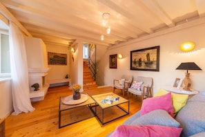 A cozy living room with wooden floors, a staircase, a sofa with colorful cushions, and a fireplace.