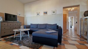 Un salon avec un canapé gris, une petite table blanche, un téléviseur posé sur un meuble et un tapis à motifs.