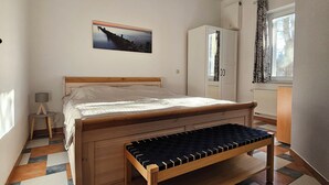 Une chambre à coucher avec un lit, un banc, un tableau, une armoire et une fenêtre avec des rideaux.