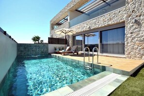A modern pool area with clear water, a stone wall, and a lounge with a table and chairs. - Villa 101 A Rhodes – Luxury Stone-Built Retreat with Pools & Panoramic Views (Ixia)