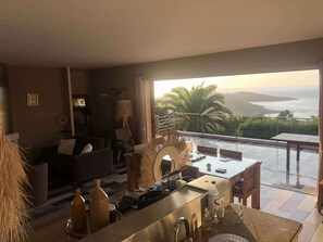 Un salon moderne comprenant un canapé, une table à manger et un coin cuisine avec un évier et un plan de travail. Une grande fenêtre offre une vue sur des palmiers et une étendue d’eau.