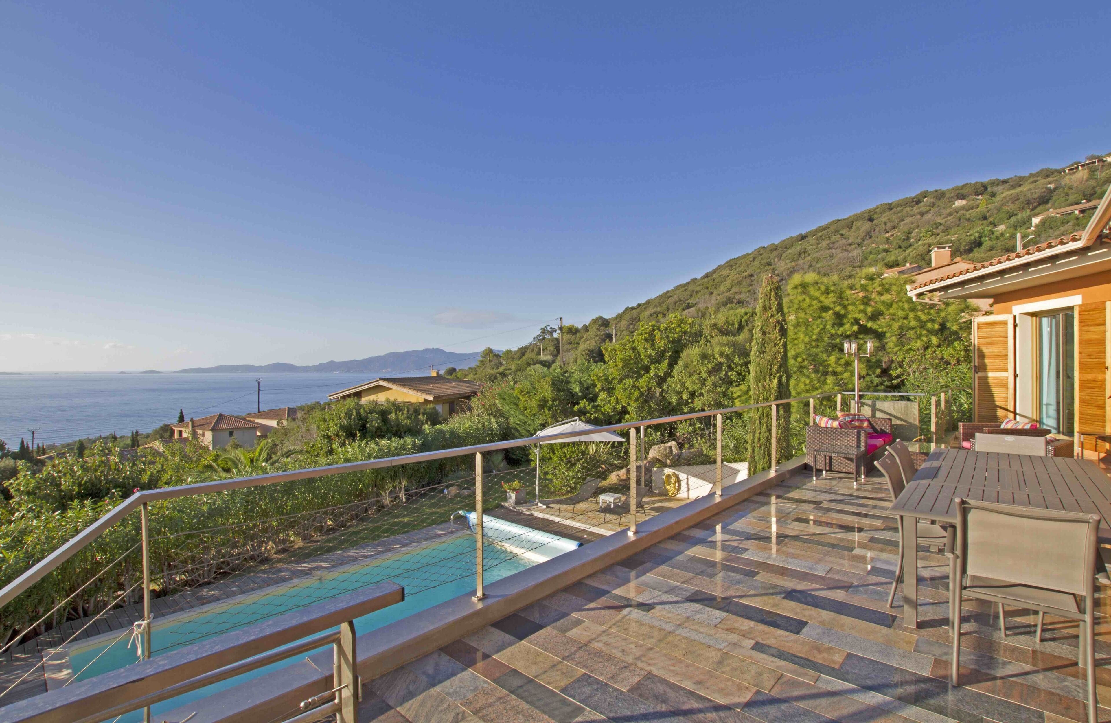 A rooftop pool area with a view of the sea, outdoor furniture, and a stone-tiled deck.
