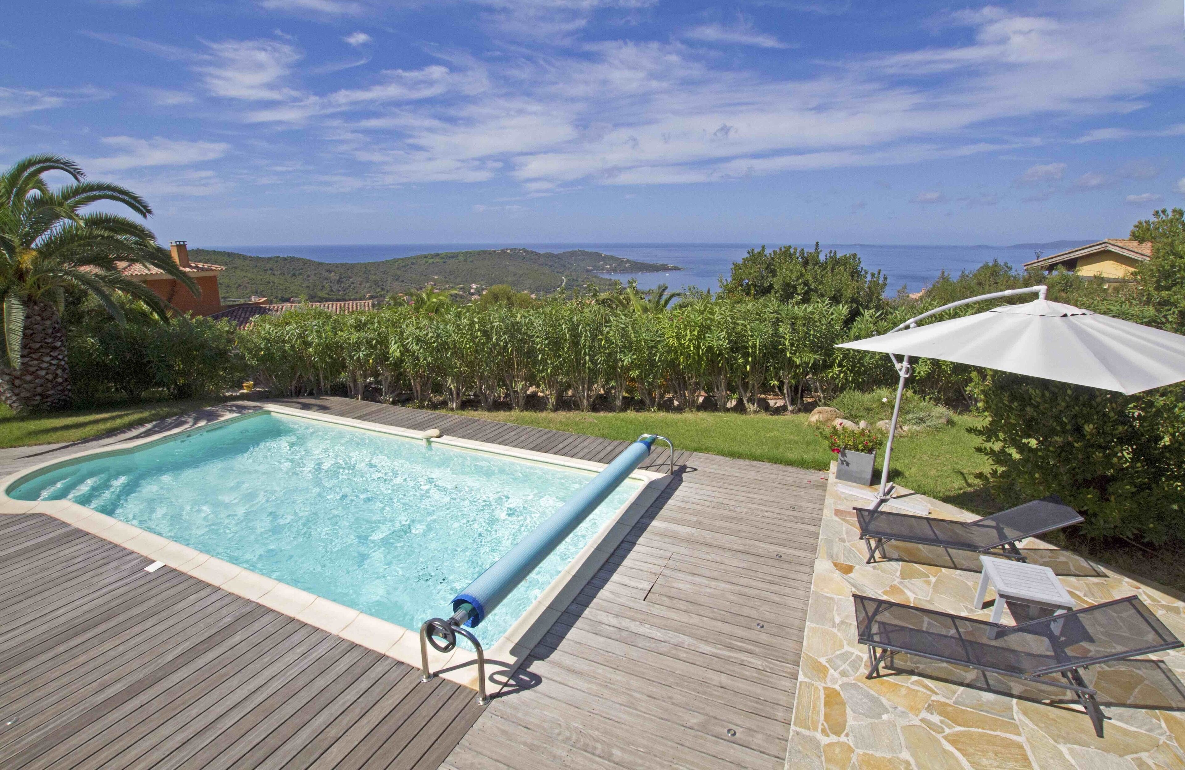 A swimming pool with a wooden deck and lounge chairs, overlooking a scenic landscape with hills and a view of the sea.