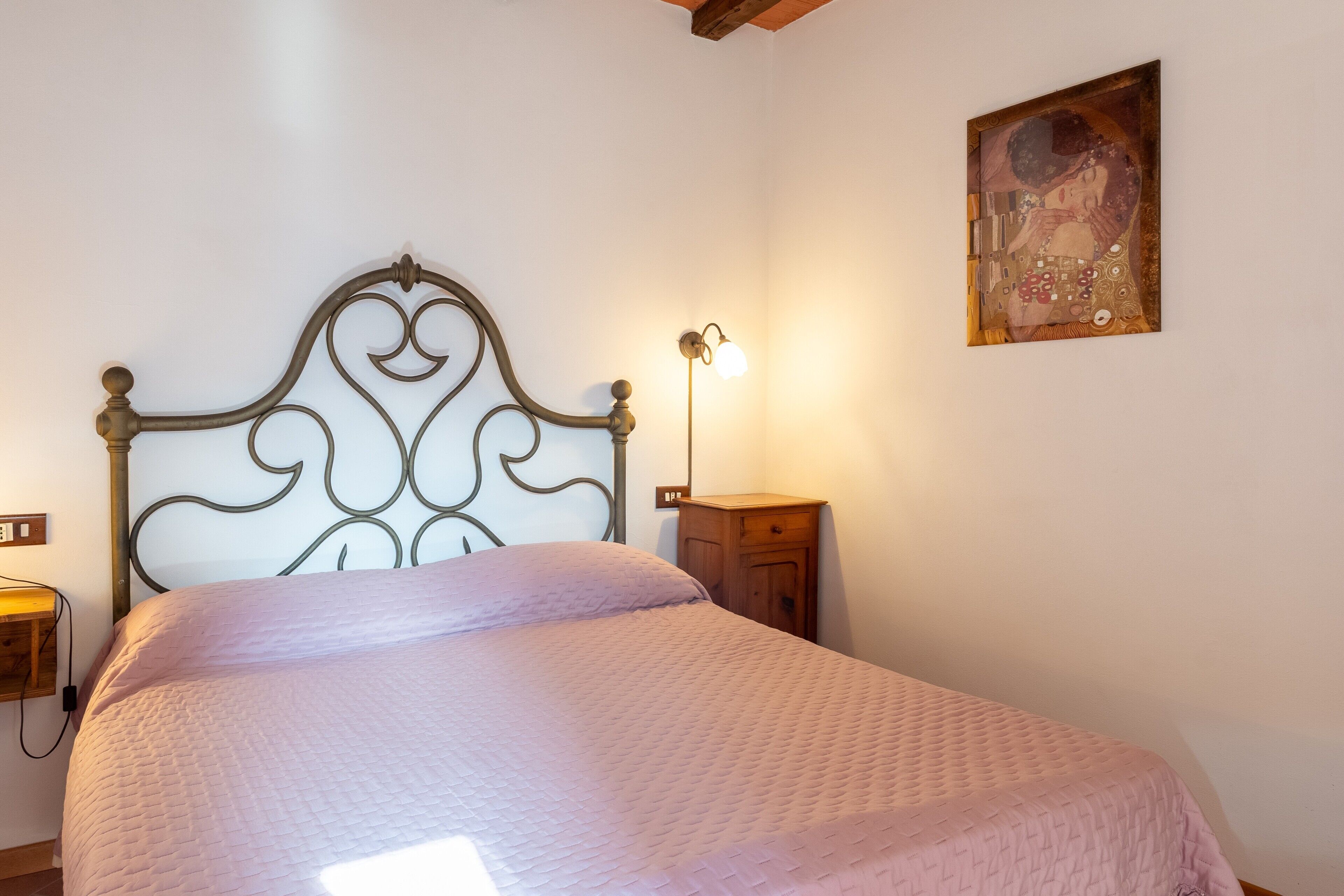 Een netjes opgemaakt bed met een gewatteerde spreidek, een houten nachtkastje met een lamp, en een schilderij aan de muur.