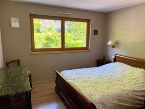 A bedroom with a bed, a desk, a window with a view of greenery, a wall-mounted lamp, and a small bedside table. - Gîte de l'Api de Broque (Fréland)