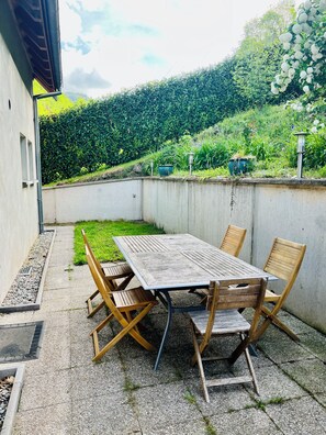 A patio with a table and chairs, a concrete wall, and a garden with plants and a hedge in the background. - Gîte de l'Api de Broque (Fréland)