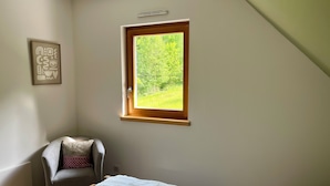 A bedroom with a sloped ceiling, a window with a view of greenery, a chair, and a bed with a white blanket. - Gîte de l'Api de Broque (Fréland)