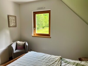 A bedroom with a sloped ceiling, a window with a view of greenery, a chair, and a bed with a white blanket. - Gîte de l'Api de Broque (Fréland)