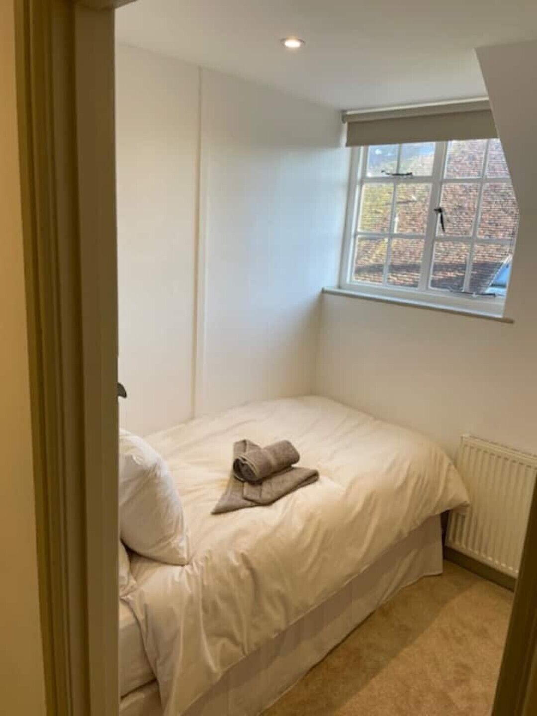 A small, neatly made bed in a room with a window, a radiator, and a door.