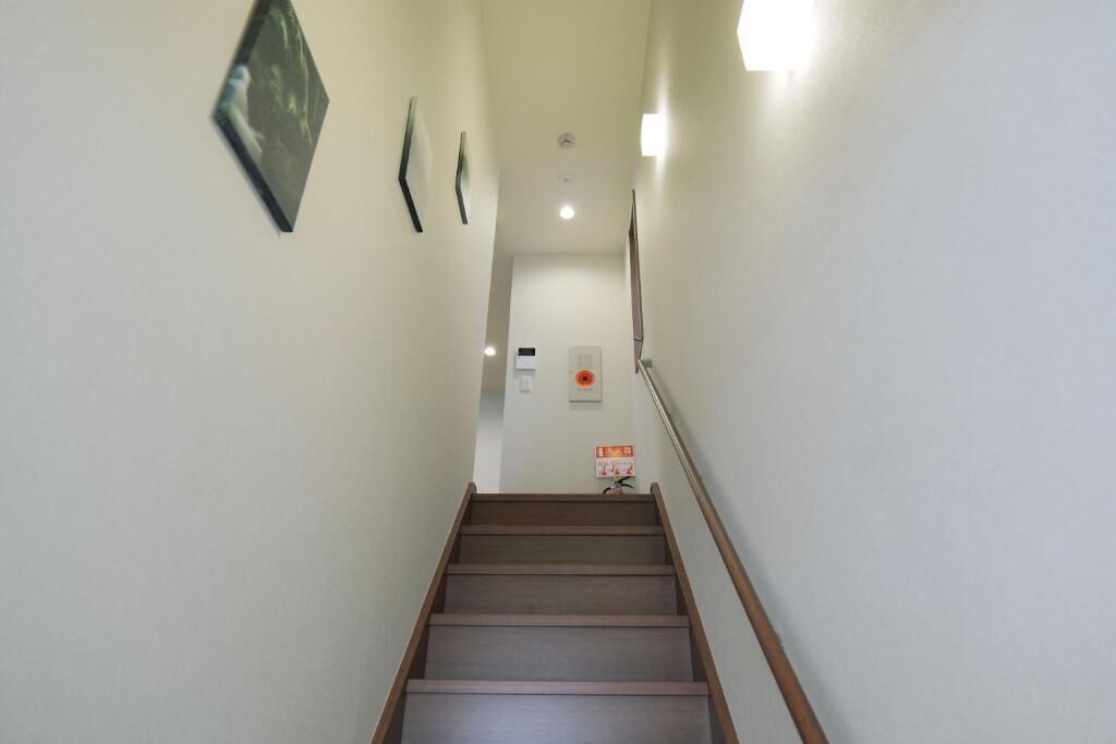 Un escalier étroit avec des marches en bois et une rampe, menant à un palier où se trouvent un luminaire et un panneau indicateur.