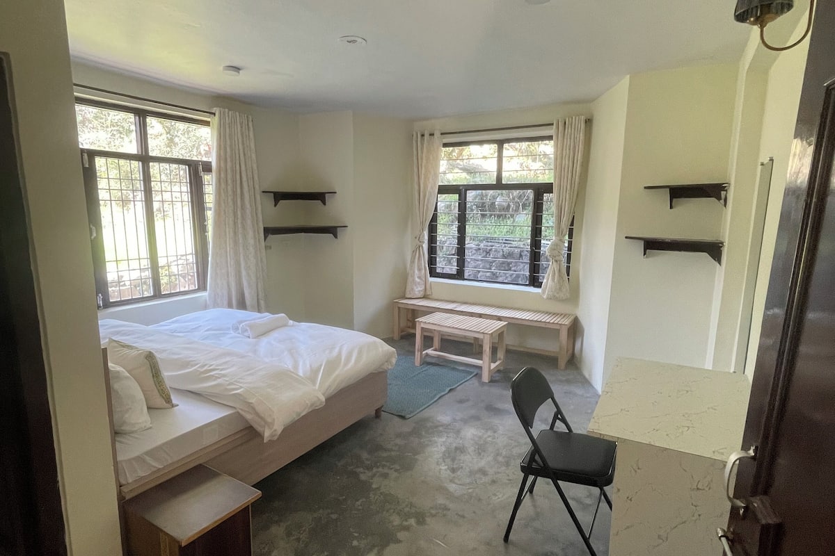 Een slaapkamer met een bed, een bureau en een stoel. Er zijn ramen met gordijnen, een bankje en planken aan de muren.