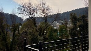 Ein Balkon mit Metallgeländer, der auf eine gebirgige Landschaft mit Bäumen und Gebäuden in der Ferne blickt.