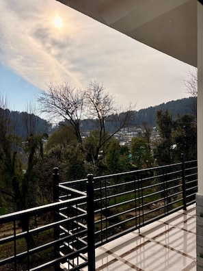 Un balcon doté d’une balustrade en métal, d’où l’on peut admirer un paysage montagneux avec des arbres et des bâtiments visibles au loin.