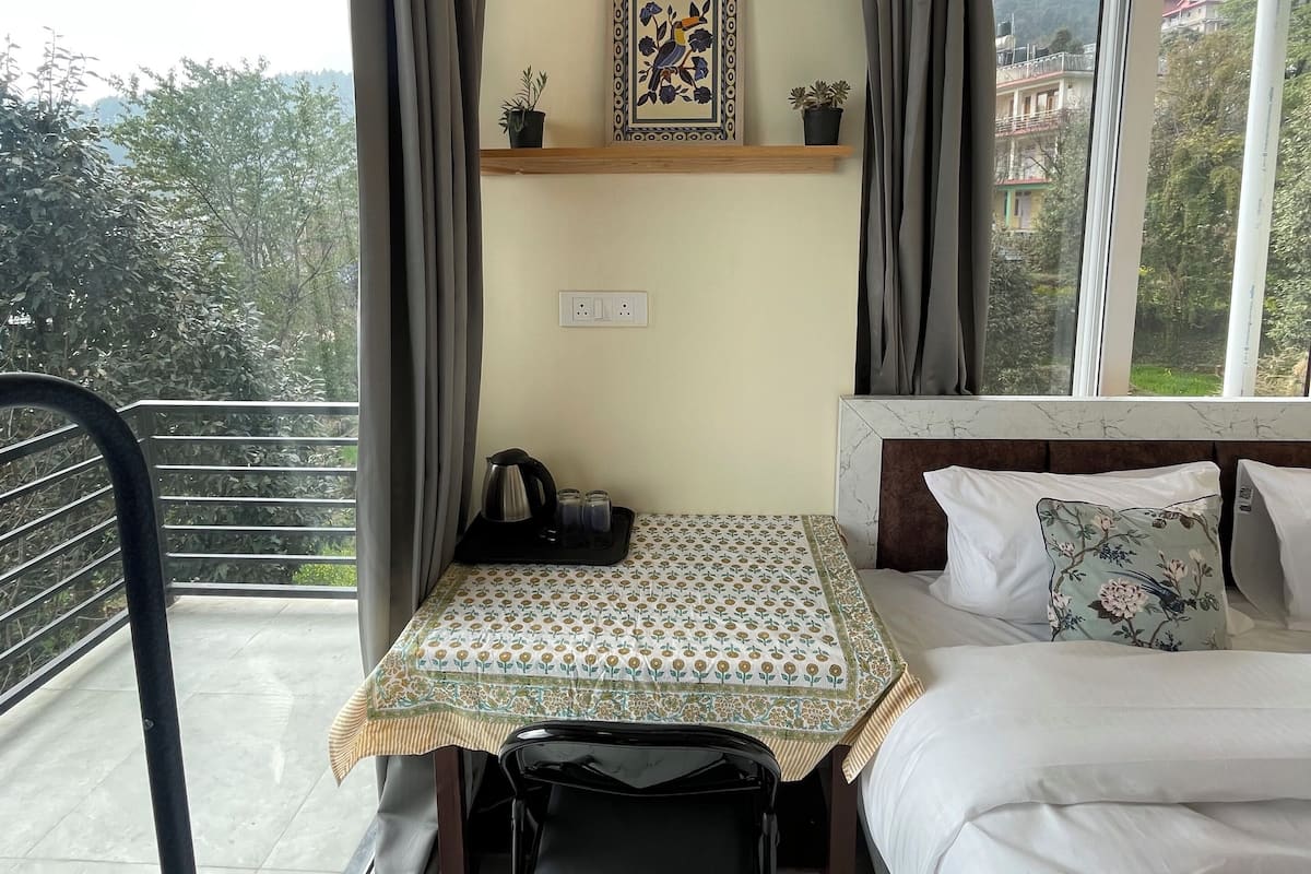 Een slaapkamer met een bed, een klein tafeltje met een dienblad, een stoel, een raam met gordijnen, een plank met een ingelijst foto en uitzicht op buiten.