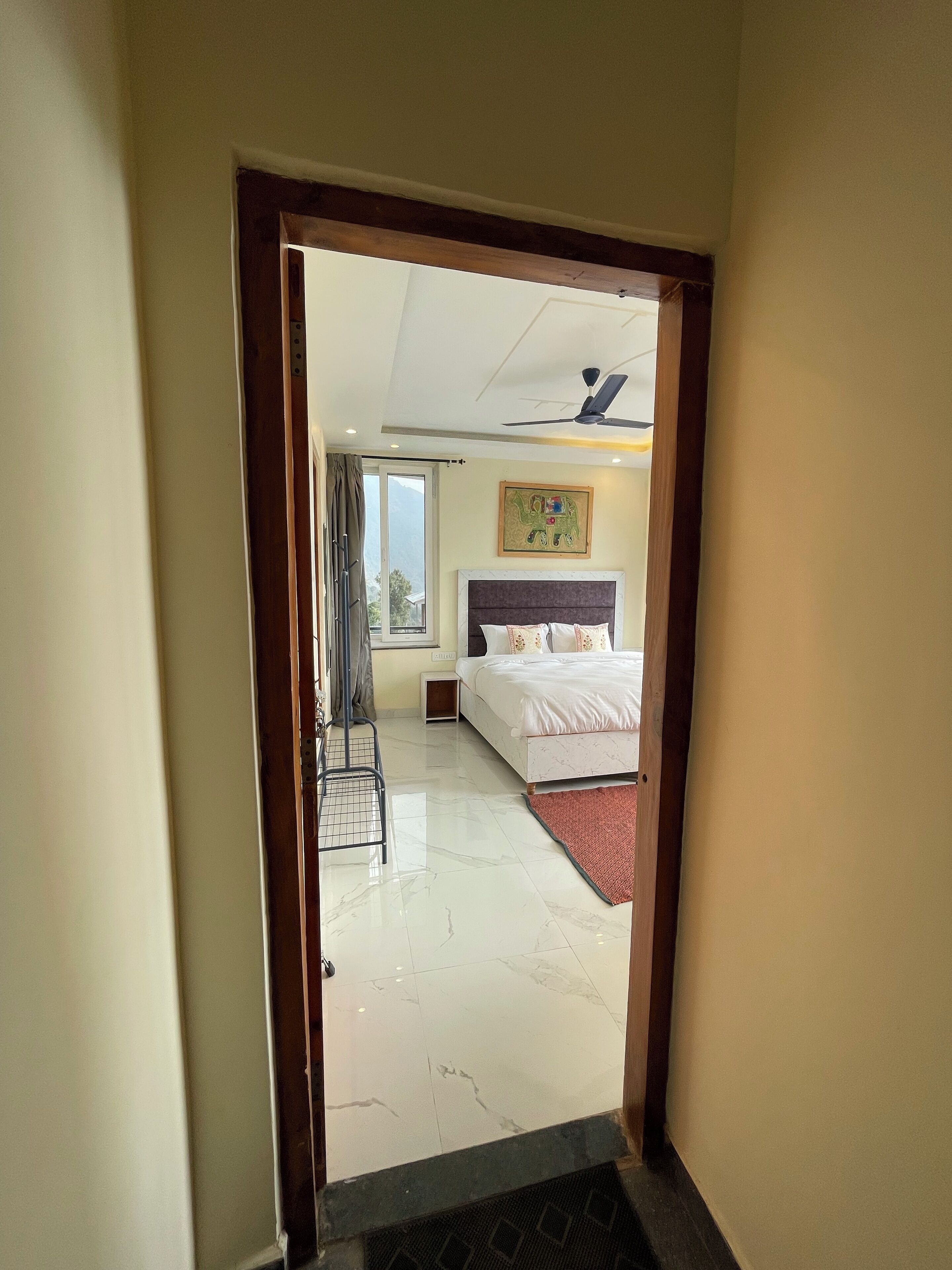 A hotel room with a bed, a ceiling fan, a window with curtains, and a painting on the wall.
