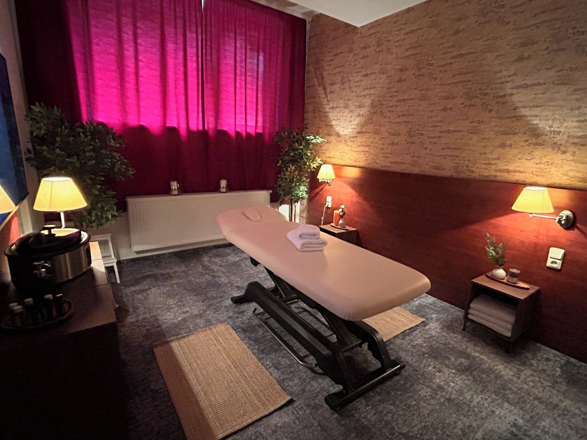 Un cuarto de terapia de masajes con una camilla, dos mesitas de noche con lámparas, un radiador y una cortina roja.