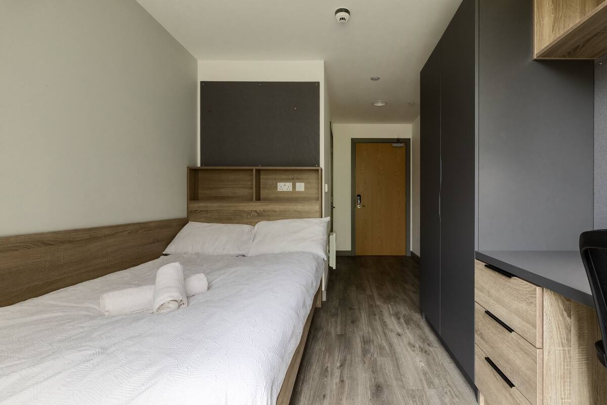 A modern hotel room with a wooden headboard, a large bed with white linens, a dark grey wardrobe, and a wooden floor.