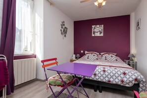 A bedroom with a bed, a purple table, a chair, a radiator, a window with curtains, a wall decoration, and a nightstand with a lamp.