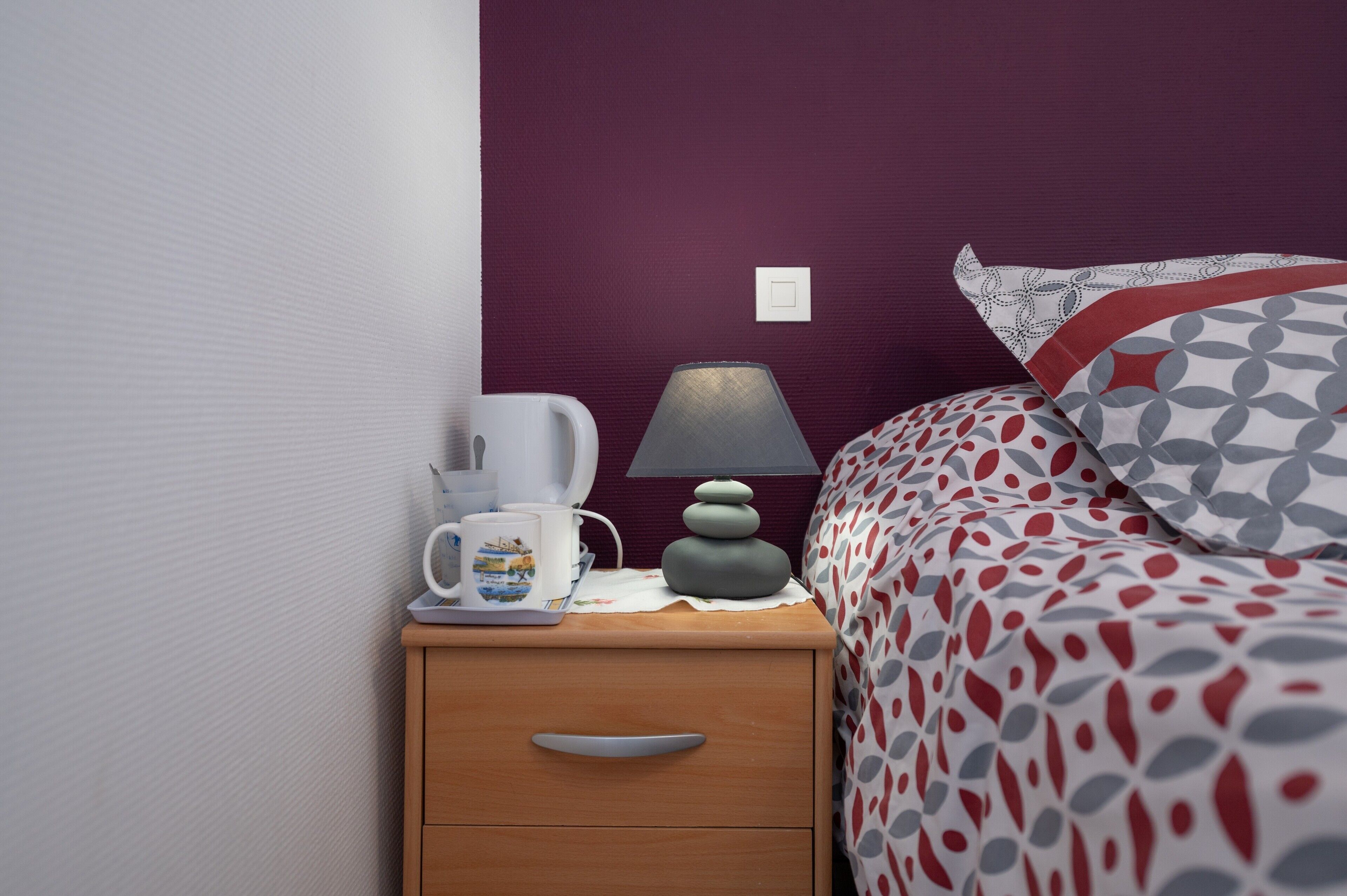Uma cama bem arrumada com roupa de cama estampada, uma luminária de cabeceira, uma cafeteira e uma gaveta pequena.