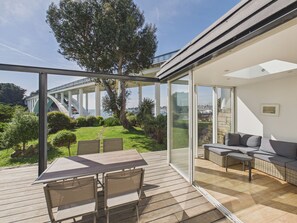 A modern outdoor patio with a dining table and chairs, a glass-enclosed seating area, and a view of a bridge and greenery. - House for 8 with garden and river view (La Trinité-sur-Mer)