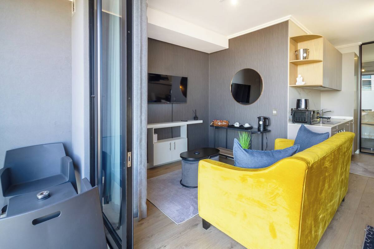 A modern living room with a yellow sofa, a flat-screen TV, and a kitchen area with a microwave and coffee machine.