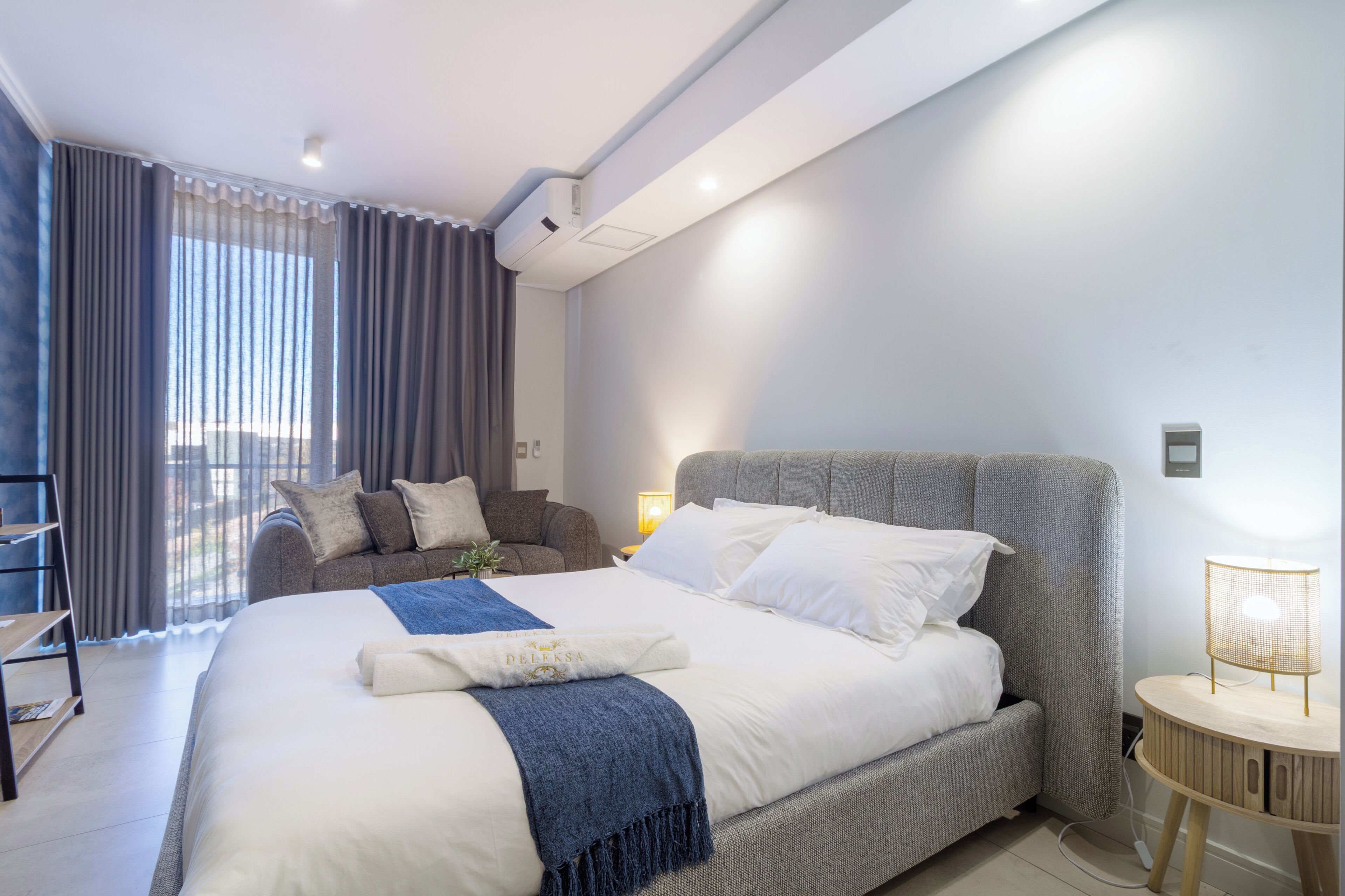 Moderni hotellihuone, jossa on suuri sänky, yöpöytä lampun kanssa, sohva ja näkymä ulos ikkunasta.