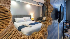 A modern hotel room with a large bed, a television, a wall-mounted air conditioner, and a wicker chair. - Lux Ellipse Galelileo (Midrand)