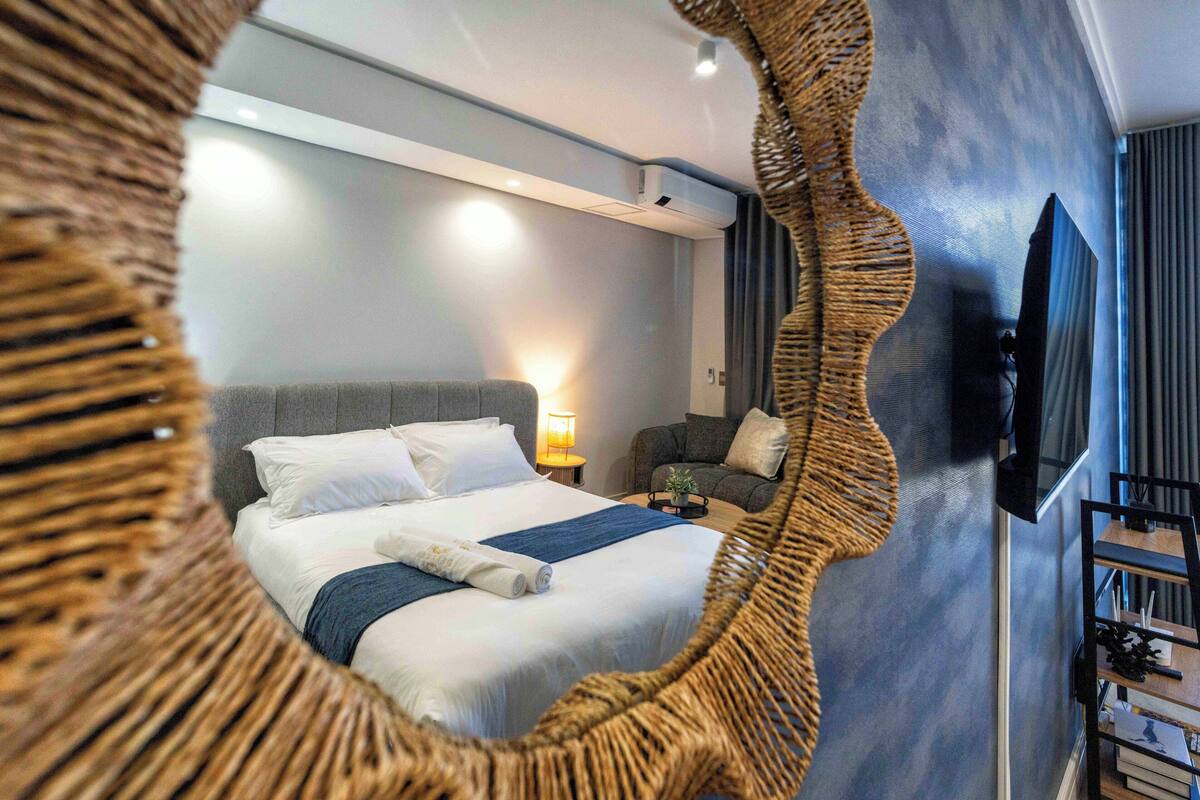 A modern hotel room with a large bed, a television, a wall-mounted air conditioner, and a wicker chair.
