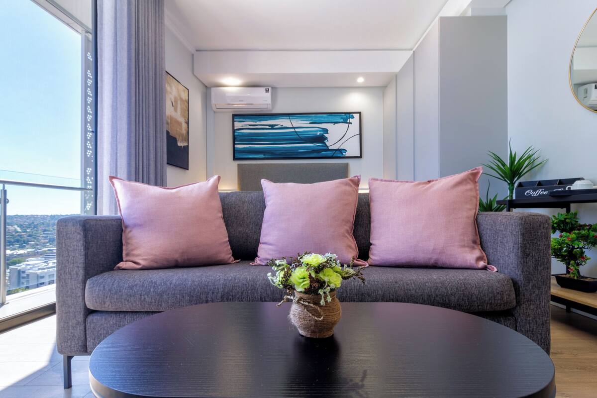 A modern living room with a grey sofa, pink cushions, a round black table, and a vase with green plants.