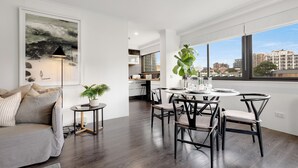 Ein modernes Wohnzimmer mit einer Couch, einem Esstisch mit Stühlen, einem kleinen Beistelltisch mit Pflanze und einem großen Fenster mit Blick auf die Stadt.